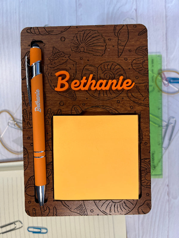 Wooden notepad with 'Bethanie' branding, orange pen, and sticky notes on a white surface. From She Shed Designs
