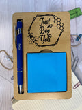 Wooden desk organizer with a pen, sticky notes, and decorative text on a light surface.