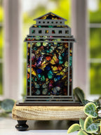 Decorative lantern with butterfly design on a wooden base in a garden setting. From She Shed Designs