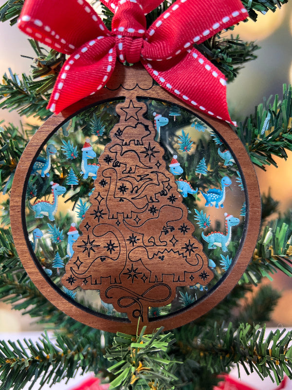 Wooden ornament with intricate dinosaur design on a Christmas tree. Layered behind with Dinosaurs wearing Santa hats. From She Shed Designs