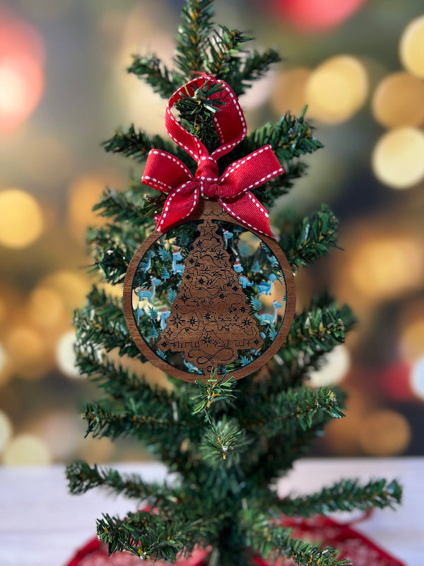 Small decorated Christmas tree with a red ribbon Wooden ornament with intricate dinosaur design on a Christmas tree. Layered behind with Dinosaurs wearing Santa hats. From She Shed Designs