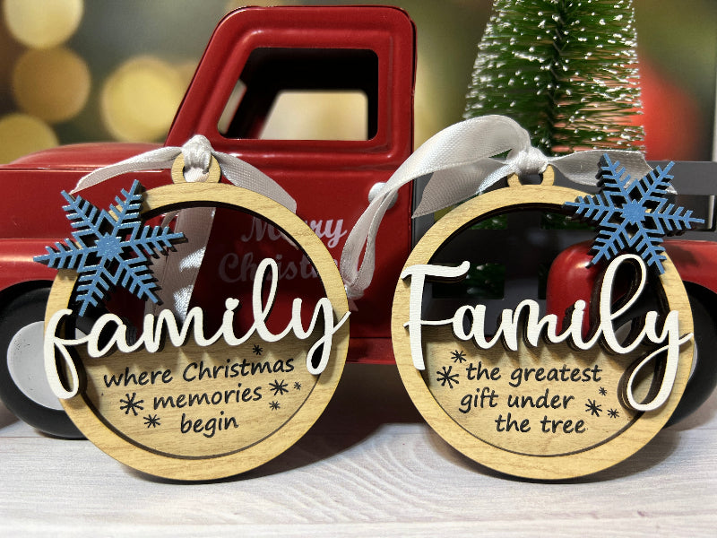 Two Christmas ornaments with 'family' text and snowflake designs in front of a red truck and Christmas tree. From She Shed Designs
