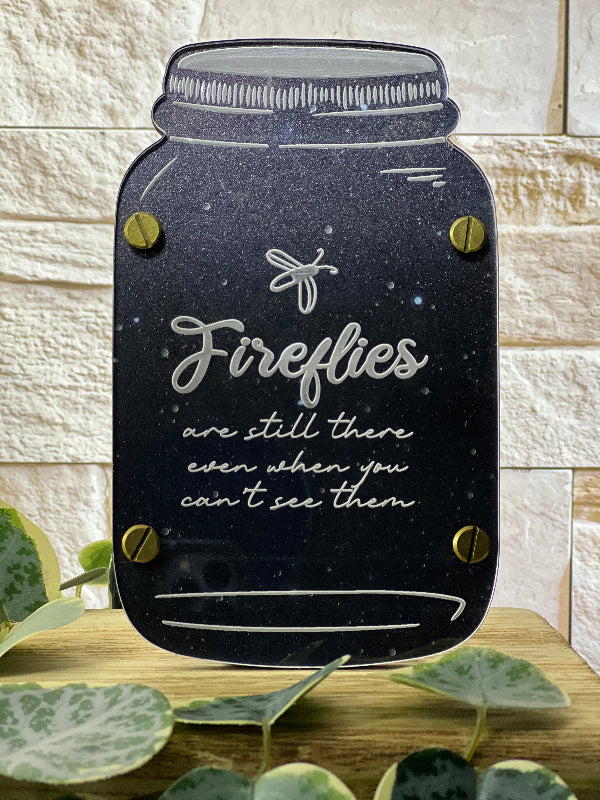 Decorative sign shaped like a mason jar with 'Fireflies are still there even when you can't see them' text on a wooden surface. From She Shed Designs
