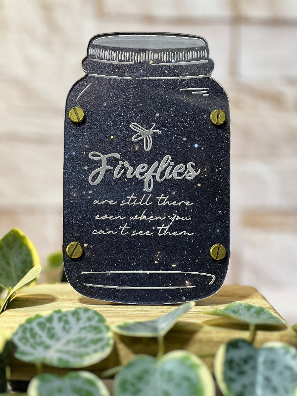 Decorative sign shaped like a mason jar with 'Fireflies are still there even when you can't see them' text, placed on a surface with green leaves. From She Shed Designs