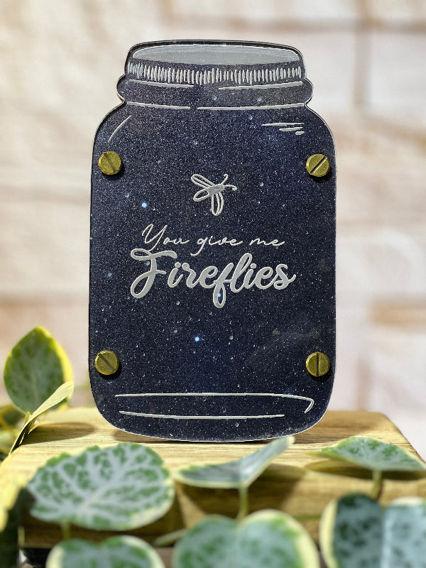 Decorative sign shaped like a mason jar with text 'You give me fireflies' on a wooden surface with leaves. From She Shed Designs