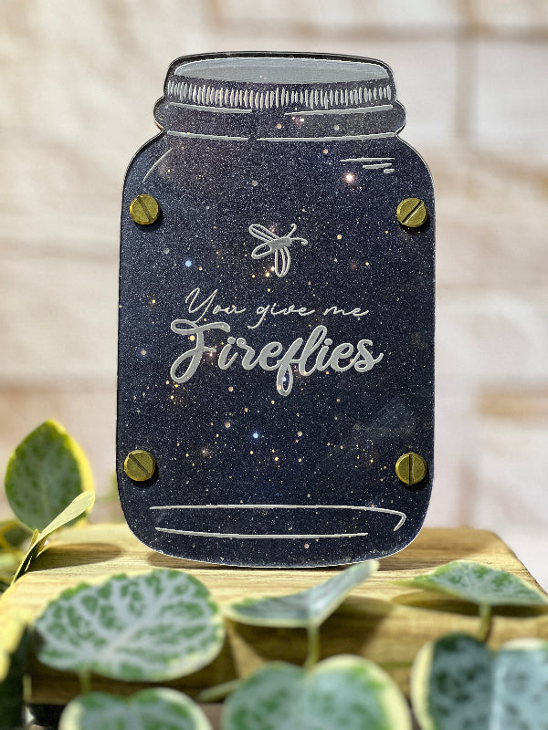 Decorative mason jar with 'You give me fireflies' text on a wooden surface with green leaves. From She Shed Designs