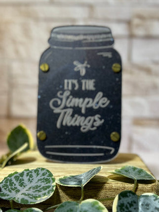 Decorative sign with 'It's the Simple Things' text on a wooden stand with green leaves. From She Shed Designs