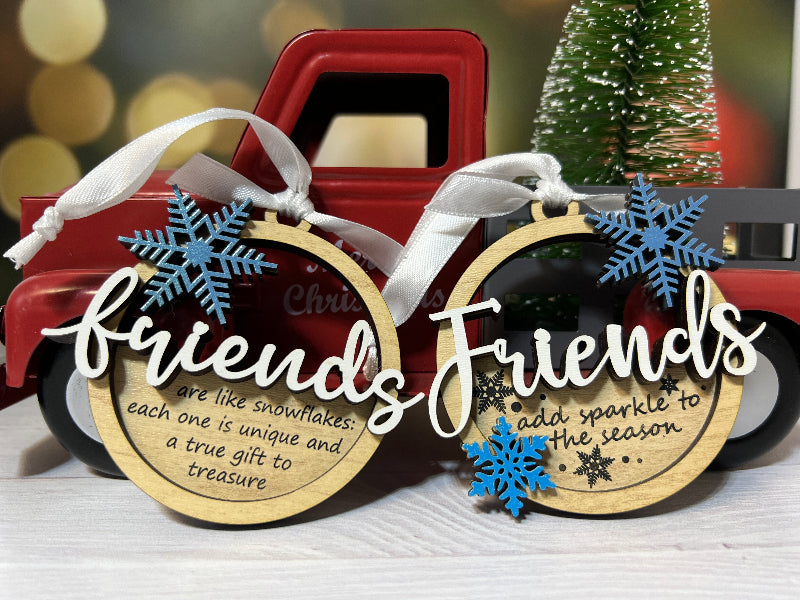 Two wooden ornaments with snowflake designs and text, placed in front of a red truck and Christmas tree. From She Shed Designs