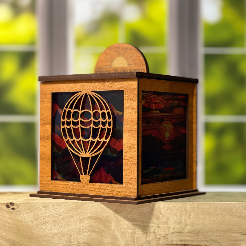 Wooden lantern with hot air balloon design on a wooden surface mountain background on 2 sides and sunset background on the other 2 sides.  half medallion on top shows sunset with words 'forever chasing sunsets'. From She Shed Designs