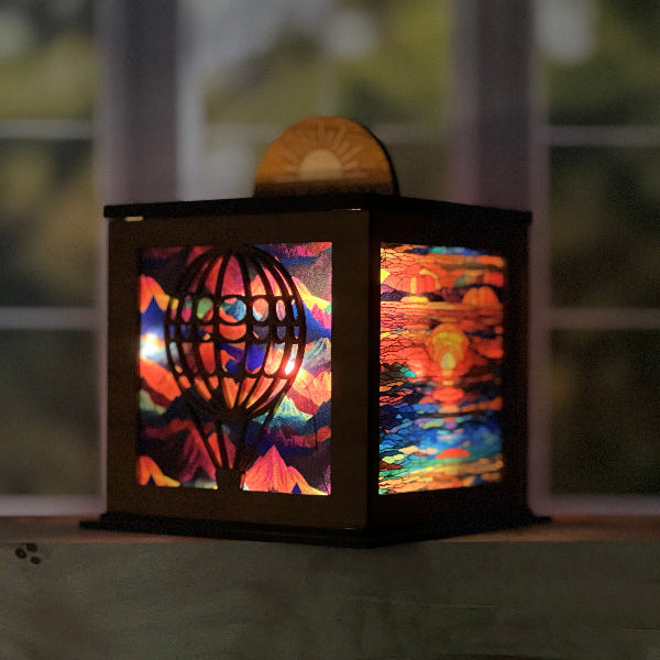 Wooden lantern with hot air balloon design on a wooden surface mountain background on 2 sides and sunset background on the other 2 sides.  half medallion on top shows sunset with words 'forever chasing sunsets'. From She Shed Designs