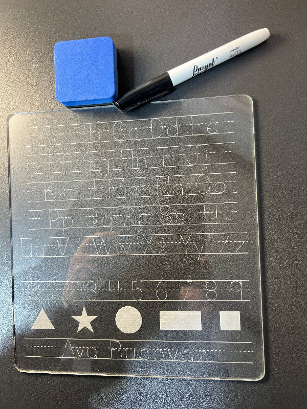 Transparent acrylic sheet with engraved letters, numbers and shapes, blue eraser, and dry erase marker on a dark surface. From She Shed Designs