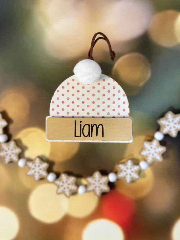 Decorative ornament with a mini snowflake hat and name tag 'Liam' against a blurred Christmas tree background. From She Shed Designs