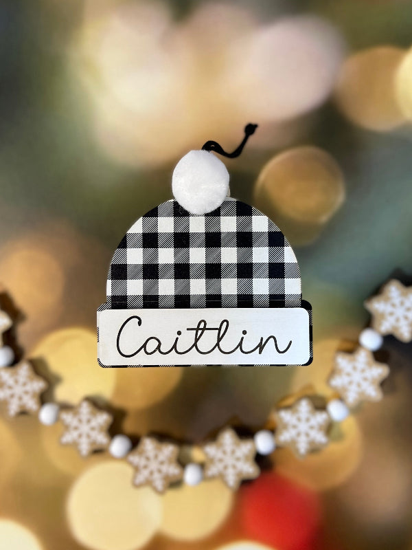 Black and white checkered hat ornament with 'Caitlin' on a blurred Christmas tree background From She Shed Designs